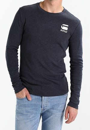 Mand iført en mørkegrå langærmet trøje med en hvid "RAW"-logo på brystet og lyseblå jeans, hånd i lommen.
