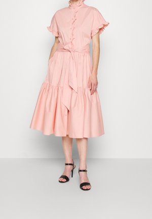 Kvinde iført en lys pink midi-kjole med flæset korte ærmer, knapper foran, bælte i taljen og sorte sandaler med høje hæle.