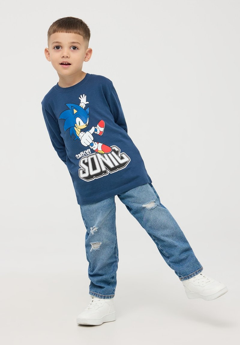 Granatowa koszula z długim rękawem z niebieską postacią z kreskówki na przodzie, połączona z podartymi jasnoniebieskimi dżinsami i białymi trampkami.