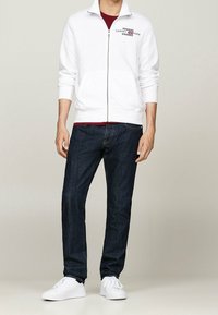 Sudadera blanca con cremallera y el logo de Tommy Hilfiger en el pecho, combinada con jeans de mezclilla oscura y zapatillas blancas. Tela suave, corte casual.