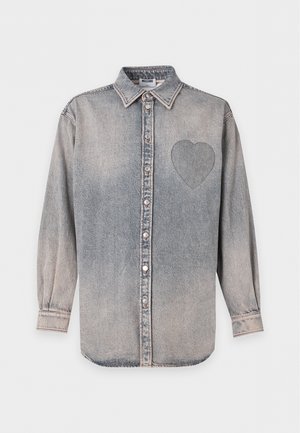 Homme portant une chemise en jean grise délavée à boutons avec un détail cœur brodé, pantalon noir avec une fermeture éclair latérale et des mocassins noirs, debout sur fond blanc.