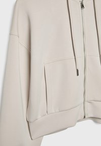 Sweat à capuche beige zippé, avec poches avant, cordon à la capuche et détails de coutures visibles sur les manches et le corps.