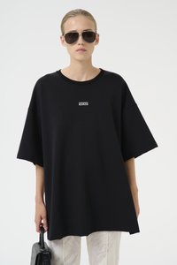 Oversized svart t-skjorte i mykt materiale, med en hvit logo på brystet, sammen med mønstrede lyse bukser og mørke solbriller.