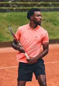 Kortermet, lys rosa polo skjorte med svart logo, kombinert med svarte shorts; holder tennisracketter på en grusbane.