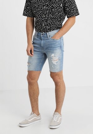 Osoba w jasnoniebieskich przetartych szortach dżinsowych, czarnej koszulce z krótkim rękawem w wzory oraz beżowych trampkach, stojąca z jedną ręką w kieszeni.