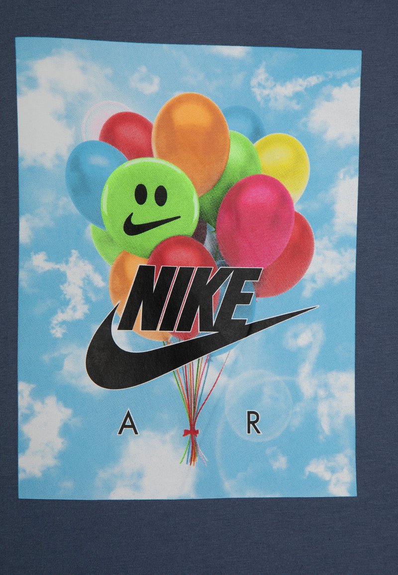 Navyblauwe T-shirt met een grafisch ontwerp van kleurrijke ballonnen met een groene glimlachende gezicht en het Nike-logo in het zwart, tegen een achtergrond van een blauwe lucht.