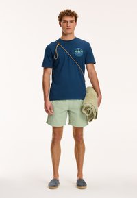 Tamsiai mėlynas marškinėliai su palmės medžio grafiką, šviesiai žali šortai, mėlynos espadrilės, laikantis suvyniotą žalią rankšluostį, yra smėlio spalvos pečių diržas.