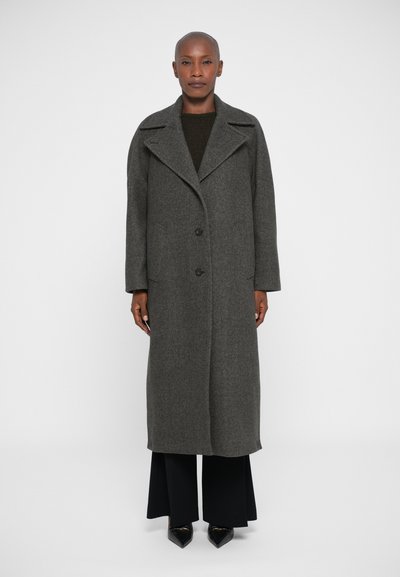 Abrigo largo de lana gris con solapa ancha, dos botones frontales y bolsillos laterales. Combinado con pantalones negros y calzado de punta.