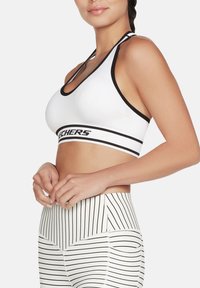 Sujetador deportivo blanco con detalles en negro y logotipo de Skechers, con textura acanalada y tirantes cruzados, combinado con leggings de tiro alto a rayas.