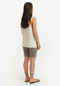 Kvinna med långt mörkt hår som bär en ärmlös beige stickad topp, taupefärgade cykelshorts och beige snörsko-flats, vänd bort från kameran.