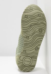 Semelle de chaussure avec des motifs ondulés et des crampons circulaires, tige en tissu vert partiellement visible, nom de la marque "WODEN" embossé au centre de la semelle.