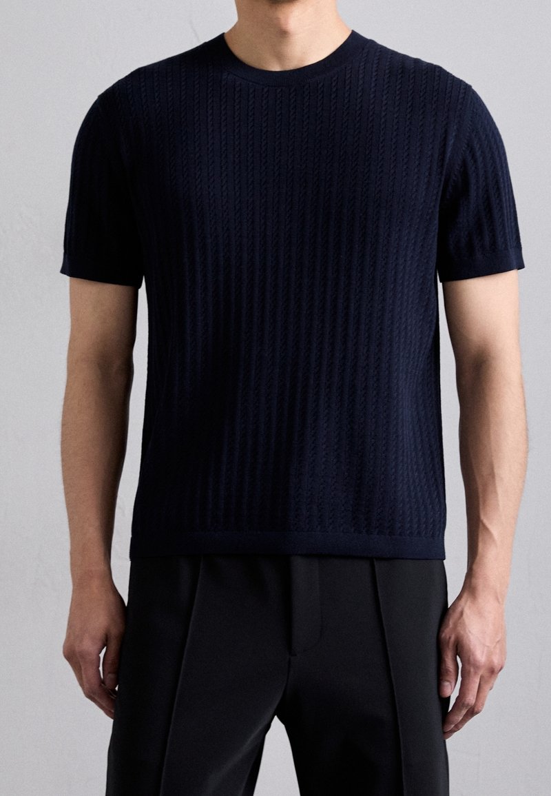 Homme portant un pull en maille texturée bleu marine à manches courtes et un pantalon noir, debout devant un fond gris clair uni.