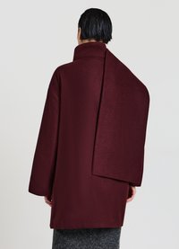 Der burgunderfarbene Wollmischmantel verfügt über einen hohen Kragen, eine übergroße Silhouette und einzigartige asymmetrische Rückenteil-Details. Glatte, strukturierte Oberfläche.