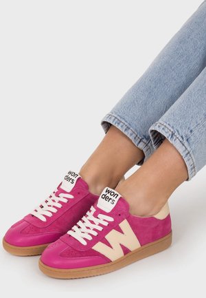 Persona con vaqueros azul claro y zapatillas deportivas rosa brillante con logotipo "W" blanco, cordones blancos y suelas color beige, sentada sobre un fondo claro.