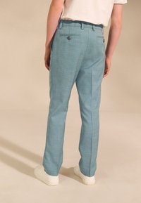 Hellblaue, strukturierte formelle Hose mit maßgeschneiderter Passform, ausgestattet mit zwei Gesäßtaschen mit Knöpfen und einem glatten Bund. Weiße Schuhe sind sichtbar.