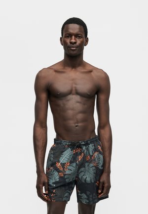 Αθλητικός μαυρισμένος άνδρας στέκεται, χωρίς πουκάμισο, φορώντας μαύρο μαγιό με πράσινα και πορτοκαλί τροπικά μοτίβα φύλλων, κοιτώντας μπροστά.
