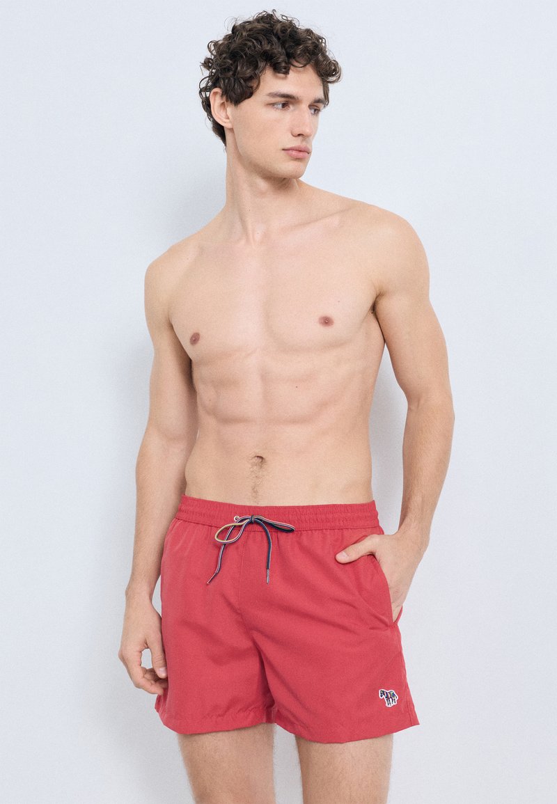 Röda badshorts med elastisk midja och blå dragsko. Har sidofickor och en liten färgglad logotyp på vänster ben.