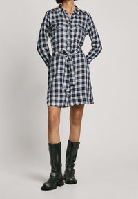 Vestito a camicia a quadri blu e bianchi con colletto, maniche lunghe, chiusura con bottoni, vita con cinturino e due tasche sul petto, abbinato a stivali neri alti fino al ginocchio.