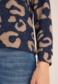 Maglione navy con motivo maculato beige, texture morbida, orlo arrotondato e maniche lunghe, abbinato a denim azzurro chiaro.