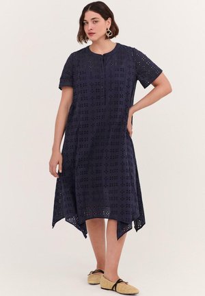 Frau trägt ein marineblaues kurzärmeliges Lochkleid mit asymmetrischem Saum und beigen flachen Schuhen mit schwarzen Riemen.