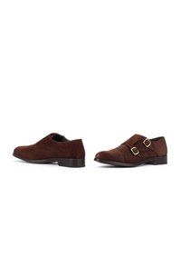 Bruna mokka kaksinkertaiset monk-strap-miesten kengät, joissa on kultaiset soljet, esillä vierekkäin valkoisella taustalla.
