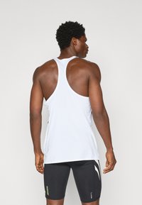 Vit ärmlös atletisk linne, i meshmaterial, med racerback-design; matchat med åtsittande svarta shorts med gröna och vita logodetaljer.