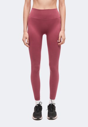 Persona che indossa leggings malva a vita alta, calzini bianchi e scarpe sportive nere, in piedi contro uno sfondo chiaro e semplice.
