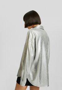 Metallisk silverfärgad långärmad skjorta med krage, avslappnad passform och knappade ärmslut, med en lätt skrynklig textur och sidorevor.