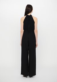 Svart jumpsuit med halterneck og knytedetalj bak. Laget av glatt stoff, har vide ben og en tettsittende midje.