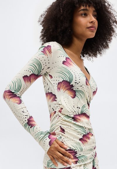 Γυναίκα με σγουρά μαλλιά που φορεί μπεζ μπλούζα με μακριά μανίκια, με μοτίβο μωβ λουλουδιών και ρίγες στο πλάι, στέκεται με το χέρι στη μέση.