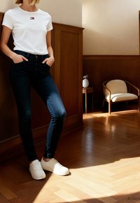Femme portant un t-shirt blanc, un jean skinny foncé et des baskets blanches, debout les mains dans les poches dans une pièce ensoleillée avec un lambris en bois.