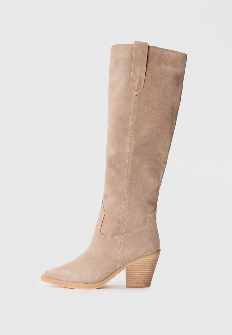 Bota de ante beige hasta la rodilla con punta afilada, costuras curvas y un tacón de madera apilada. Sin herrajes ni adornos visibles.