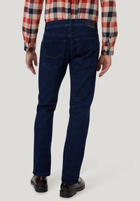 Dunkelblaue Jeans mit geradem Schnitt, ausgestattet mit Gesäßtaschen und einem Lederpatch am Bund. Kombiniert mit einem karierten Hemd und schwarzen Schuhen.