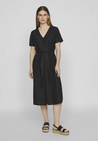 Juoda, vidutinio ilgio suknelė su trumpomis rankovėmis, V formos iškirpte, mygtukais sagstoma priekyje ir surišta juosmens srityje. Derinama su juodais espadriliais.