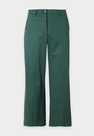 Femme debout, mains dans les poches, portant un haut rose à manches courtes, un pantalon large vert foncé et des chaussures beiges à bouts pointus.