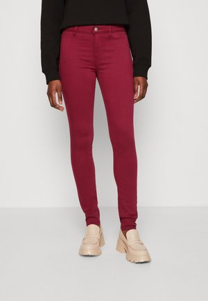Personne portant un pantalon moulant rouge foncé, un haut à manches longues noir et des mocassins à talons épais beige, debout devant un fond uni.
