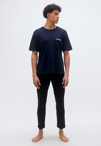 Νέος άνδρας με σγουρά μαλλιά, φοράει μπλε ναυτικό μπλουζάκι Jack & Jones και μαύρο παντελόνι στενής εφαρμογής, στέκεται ξυπόλητος και κοιτάζει προς τα αριστερά του.