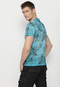 Tirkīza krāsas īsām piedurknēm krekls ar melniem ziedu un lapu rakstiem, izgatavots no viegla auduma, ar nedaudz brīvu piegriezumu un apaļu kaklaizdari.