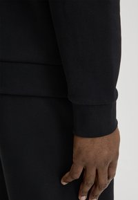 Zwarte trui met geribbelde manchetten en zoom, gestructureerde stof, nauwsluitend ontwerp, met een deel van een hand die op de taille rust.