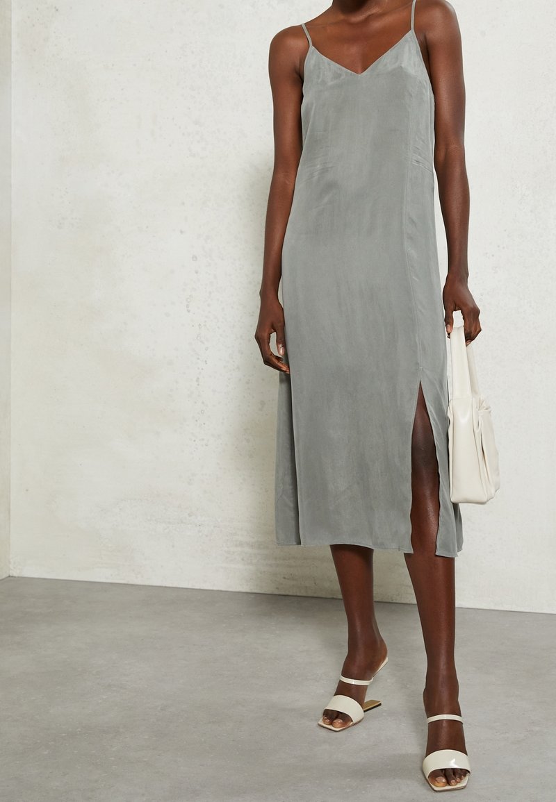 Szara, jedwabista sukienka na ramiączkach z dekoltem w serek i rozcięciem po bokach. Do tego kremowa torebka i beżowe sandały na obcasie.