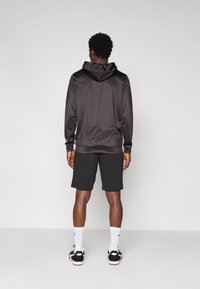 Hoodie en satin noir avec un col à cordon, associé à des shorts foncés. La tenue présente des poignets côtelés et des chaussures de sport avec des chaussettes blanches.