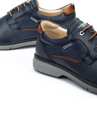 Chaussures en cuir marine avec accents marron, bout rond, semelle en caoutchouc grise et design à lacets. Présentent une étiquette logo sur le côté.