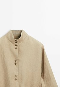 Beige liinapaita, jossa on korkea kaulus, kuuden pyöreän napin nappilista ja pitkät hihat. Sileä rakenne ja rento muotoilu.