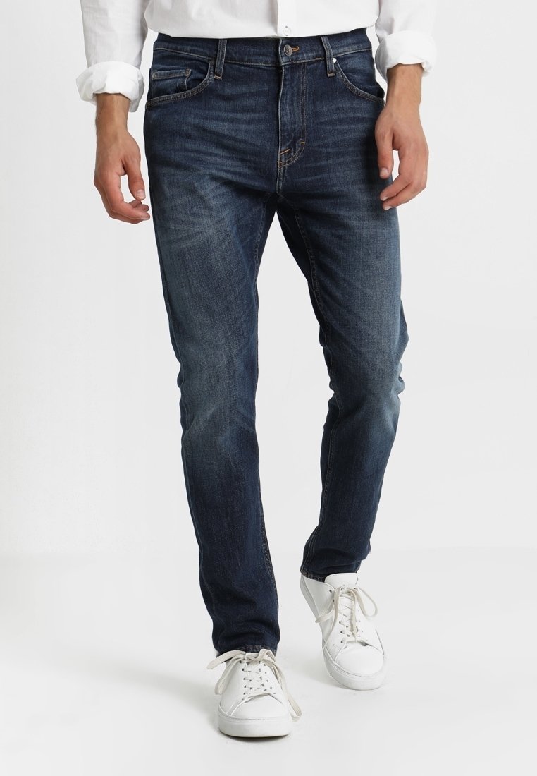 Man med mörkblå slim-fit jeans, vita sneakers och en vit långärmad skjorta med uppkavlade ärmar, stående mot en enfärgad bakgrund.