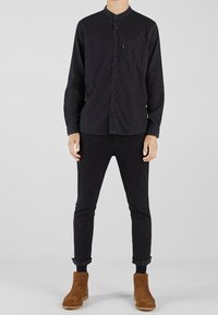 Maglietta nera a maniche lunghe con colletto alla mandarino e taschino sul petto, abbinata a jeans skinny neri e stivaletti in camoscio marrone.