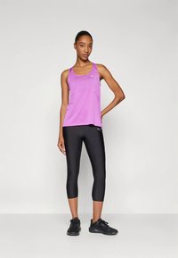 Žena stojící, oblečená v purpurovém sportovním topu bez rukávů, černých zkrácených legínách a černých sportovních botách na jednobarevném pozadí.