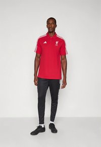 Raudonas polo marškinėlis su Liverpulio FC logotipu, juodos sportinės kelnės su baltomis šoninėmis juostelėmis ir juodi sportiniai batai. Trumpomis rankovėmis ir apykakle.