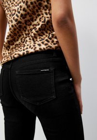 Schwarze Jeans mit einer taillierten Passform, die kontrastierende Nähte und ein kleines Logodetail an der Gesäßtasche aufweisen. Kombiniert mit einem Oberteil im Leopardenmuster.