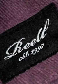 Schwarzes Stoffetikett mit weißem, gesticktem Text "Reell est. 1997" auf einem strukturierten lila Hintergrund.