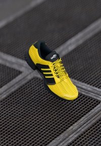 Κίτρινο αθλητικό παπούτσι με υφή, μαύρες λεπτομέρειες και τρεις μαύρες ρίγες. Διαθέτει μαλακή εσωτερική επένδυση και ευλύγιστη σόλα.